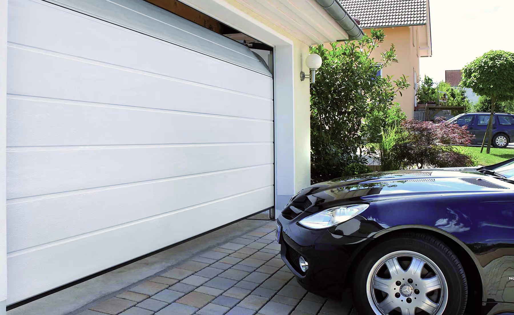 Schwarzes Auto fährt in eine weiße Garage mit geöffnetem Garagentor in einem gepflegten Wohngebiet.