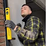 Mann in kariertem Hemd und Handschuhen hält eine Wasserwaage gegen eine Backsteinwand.