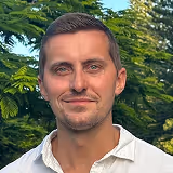 Porträt eines lächelnden Mannes mit kurzem braunem Haar und blaugrünen Augen vor grünem Laub im Freien.