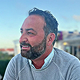 Nahaufnahme eines lächelnden Mannes mit Bart und kurzen dunklen Haaren, der nach links schaut, vor einem unscharfen Hintergrund im Freien.