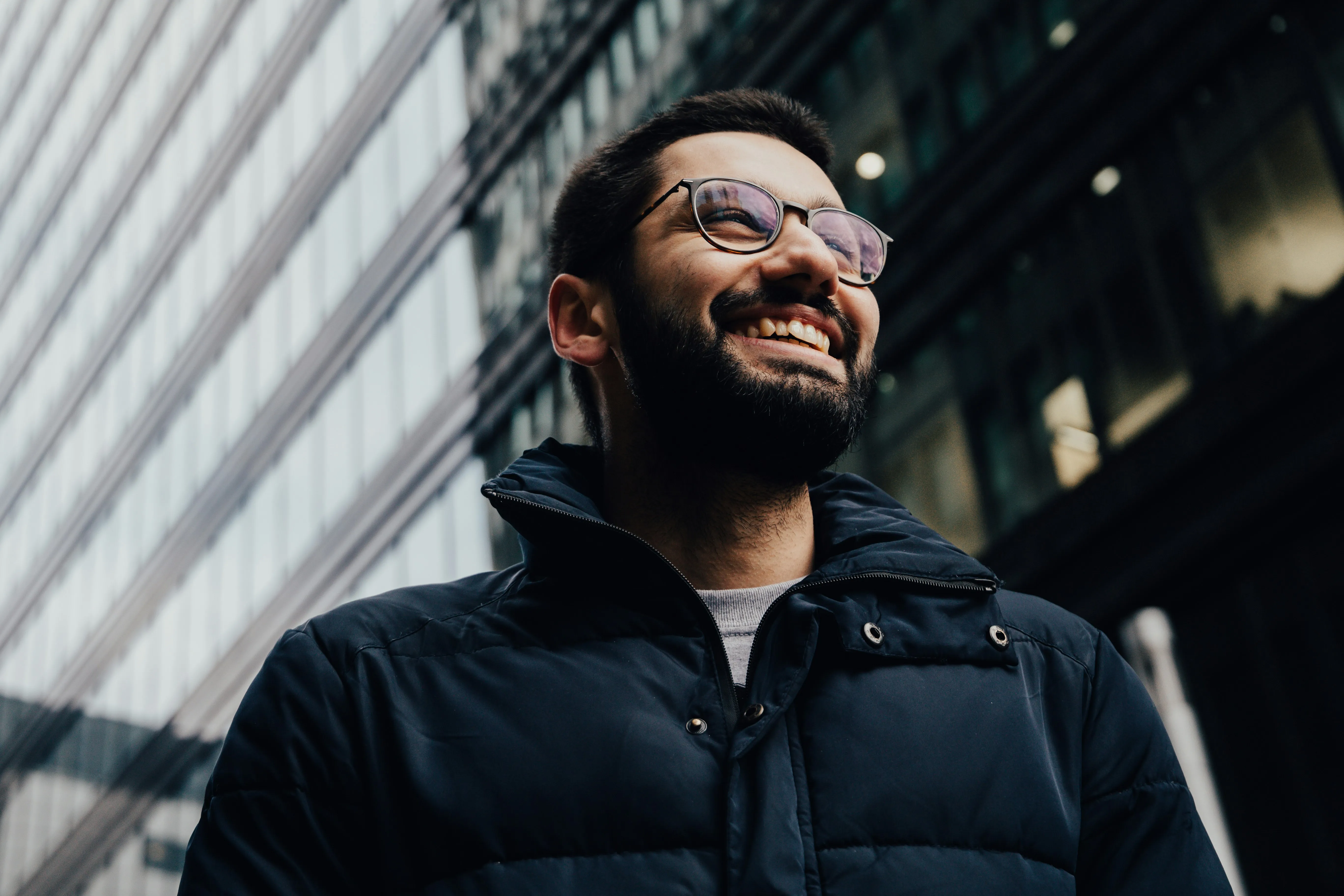 Un homme souriant avec des lunettes et une barbe portant un manteau noir devant des immeubles de bureaux modernes.