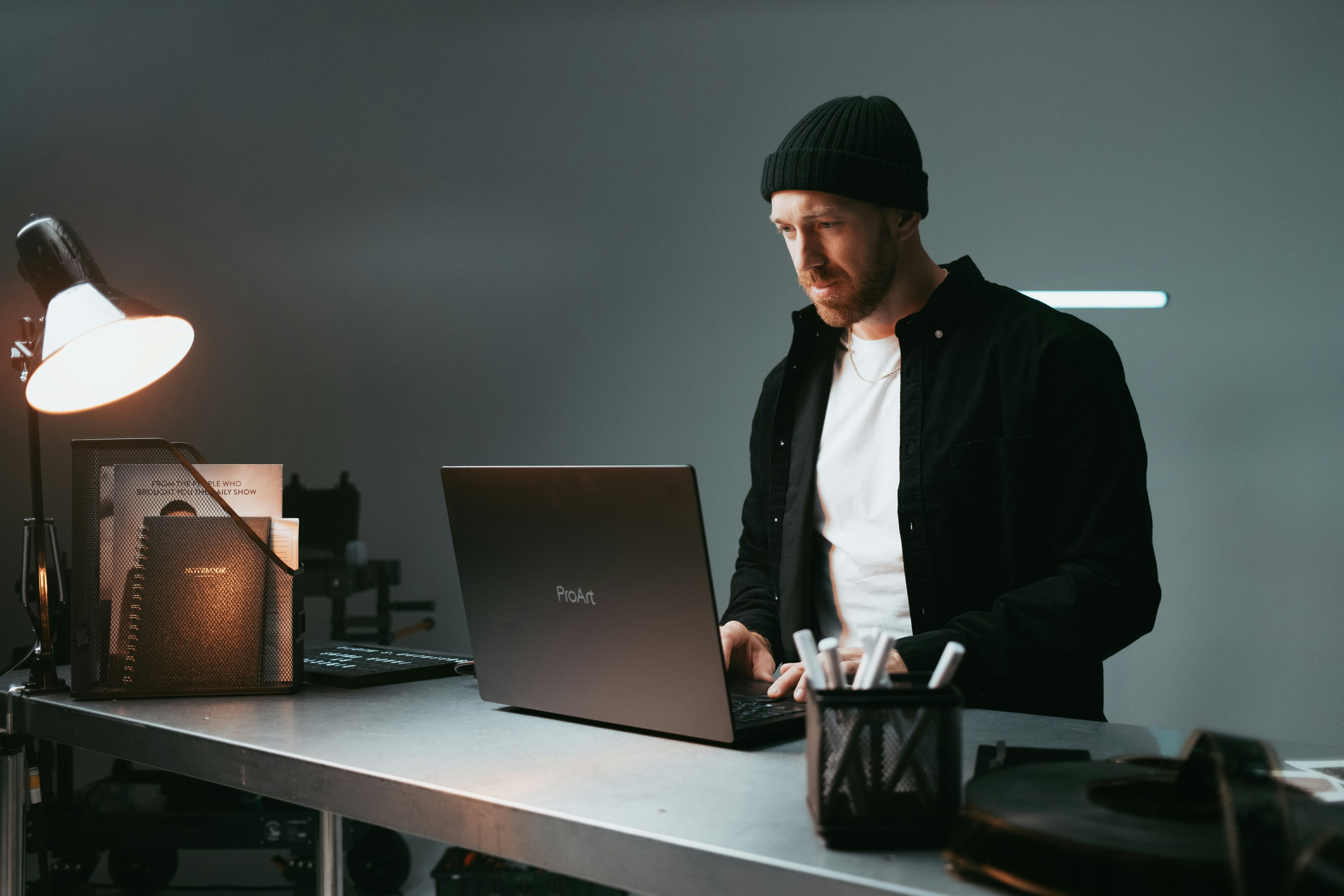 Homme portant un bonnet noir travaillant sur un ordinateur portable ProArt sur un bureau métallique avec une lampe allumée et des accessoires de bureau.
