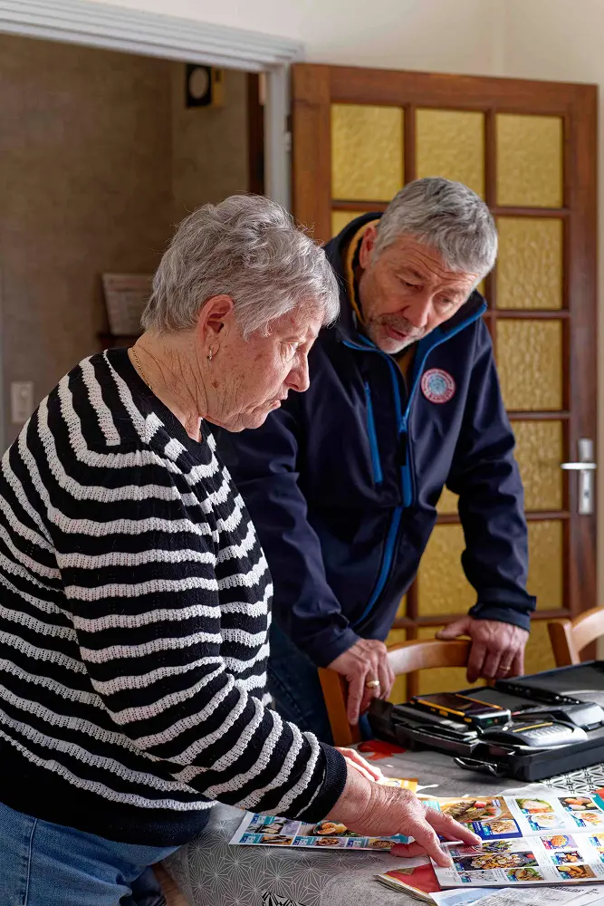 Older woman in striped sweater and older man in dark jacket looking at printed photos or postcards on a table indoors.