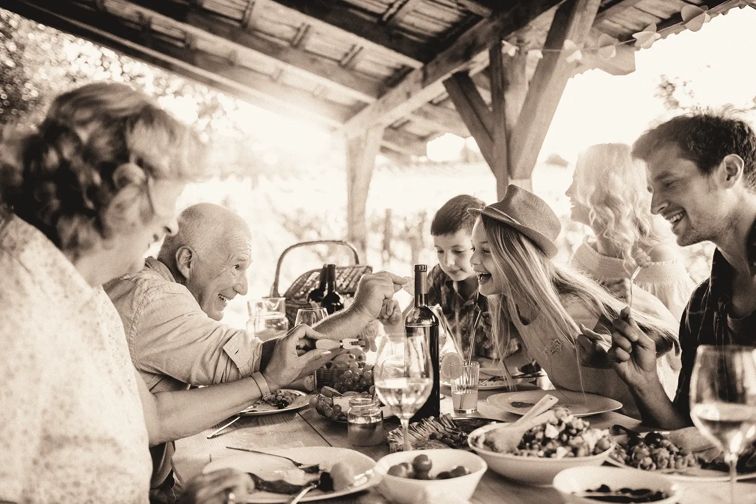 Multigenerational family laughing and sharing food while enjoying an outdoor meal under a wooden pergola.