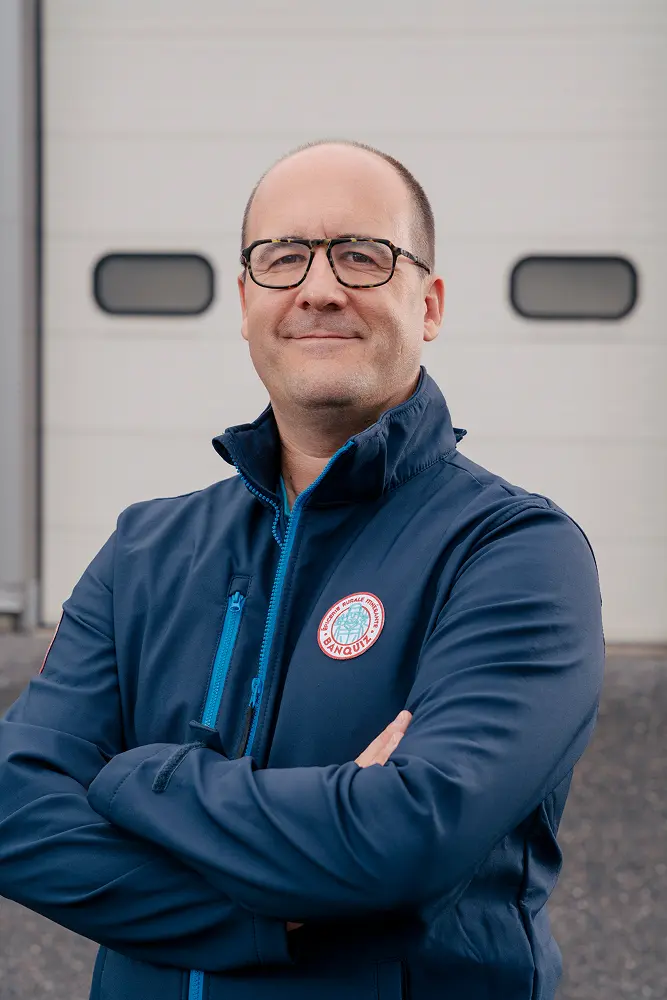 Smiling man with glasses wearing a navy blue jacket with BANQUIZ logo standing with arms crossed.