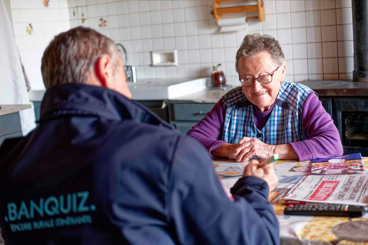 Elderly woman smiling and sitting at a kitchen table talking with a man wearing a BANQUIZ jacket.