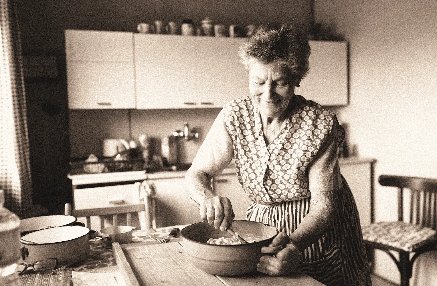 Elderly woman smiling while stirring dough in a bowl in a vintage kitchen.