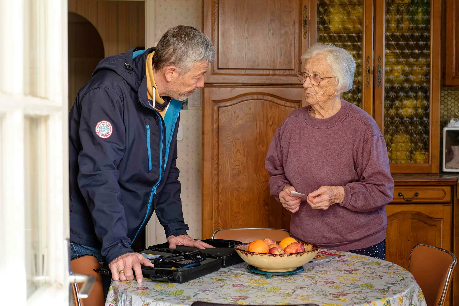 Man in a navy jacket talking to an elderly woman in a purple sweater at a kitchen table with a bowl of fruit and a toolkit.