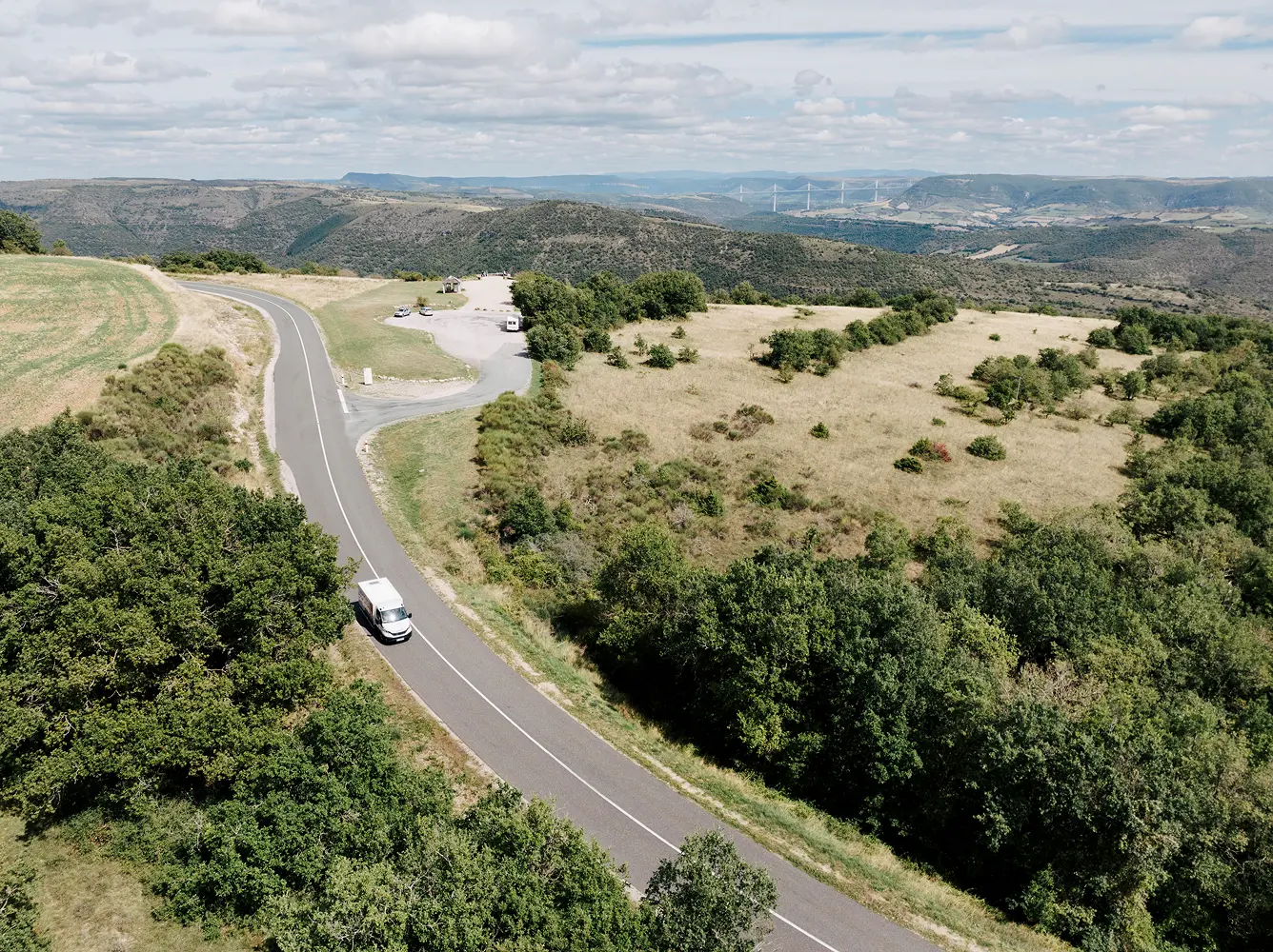 White van driving on a winding rural road surrounded by green trees and dry grass under partly cloudy skies with distant hills and a bridge visible.