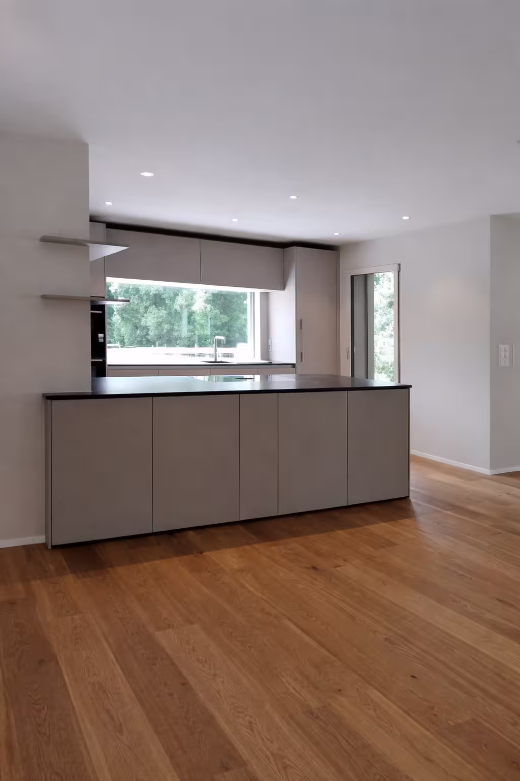 Moderne minimalistische Küche mit grauer Insel, Holzboden und großem Fenster mit Blick auf grüne Bäume.