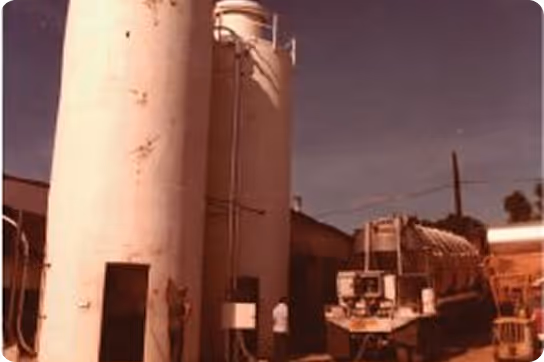 Two large vertical industrial storage tanks next to a truck with a cylindrical tank under a clear sky.