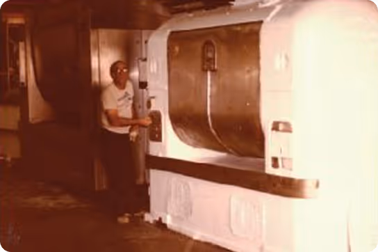 Man standing beside a large industrial machine inside a dimly lit factory or workshop.