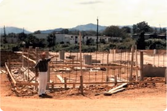 Construction site with a man wearing a red hard hat standing on dirt near wooden framing and metal rods, with mountains in the background.