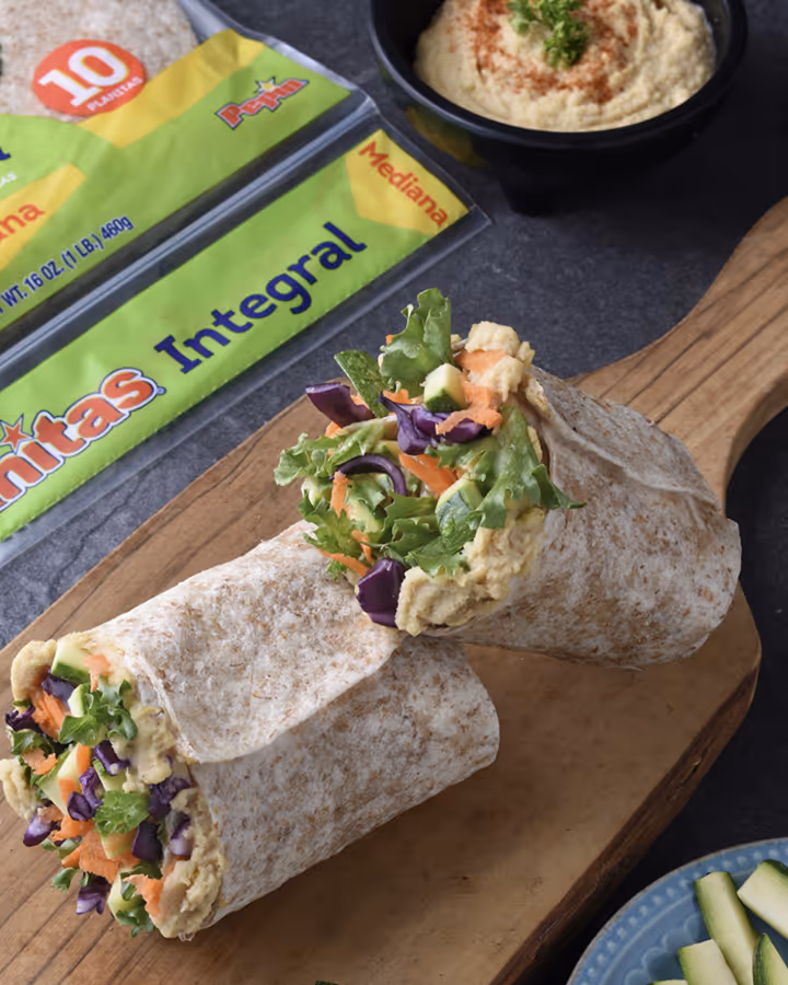 Whole wheat tortilla wrap filled with lettuce, shredded carrot, purple cabbage, and hummus on a wooden cutting board.