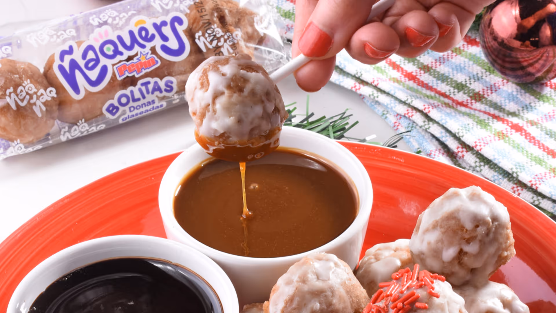 Hand with red nail polish dipping a glazed mini donut on a stick into a cup of caramel sauce, with more glazed mini donuts and a cup of chocolate sauce on a red plate.