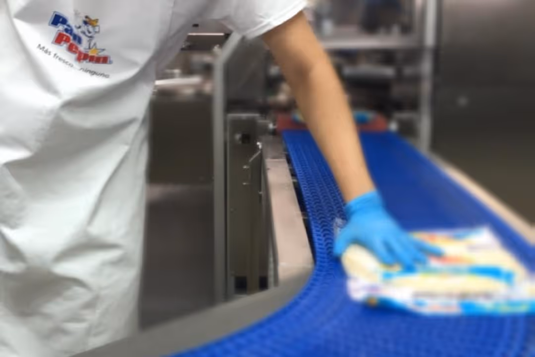 Worker wearing a white shirt with logo and blue glove placing packaged food on a blue conveyor belt in a factory.