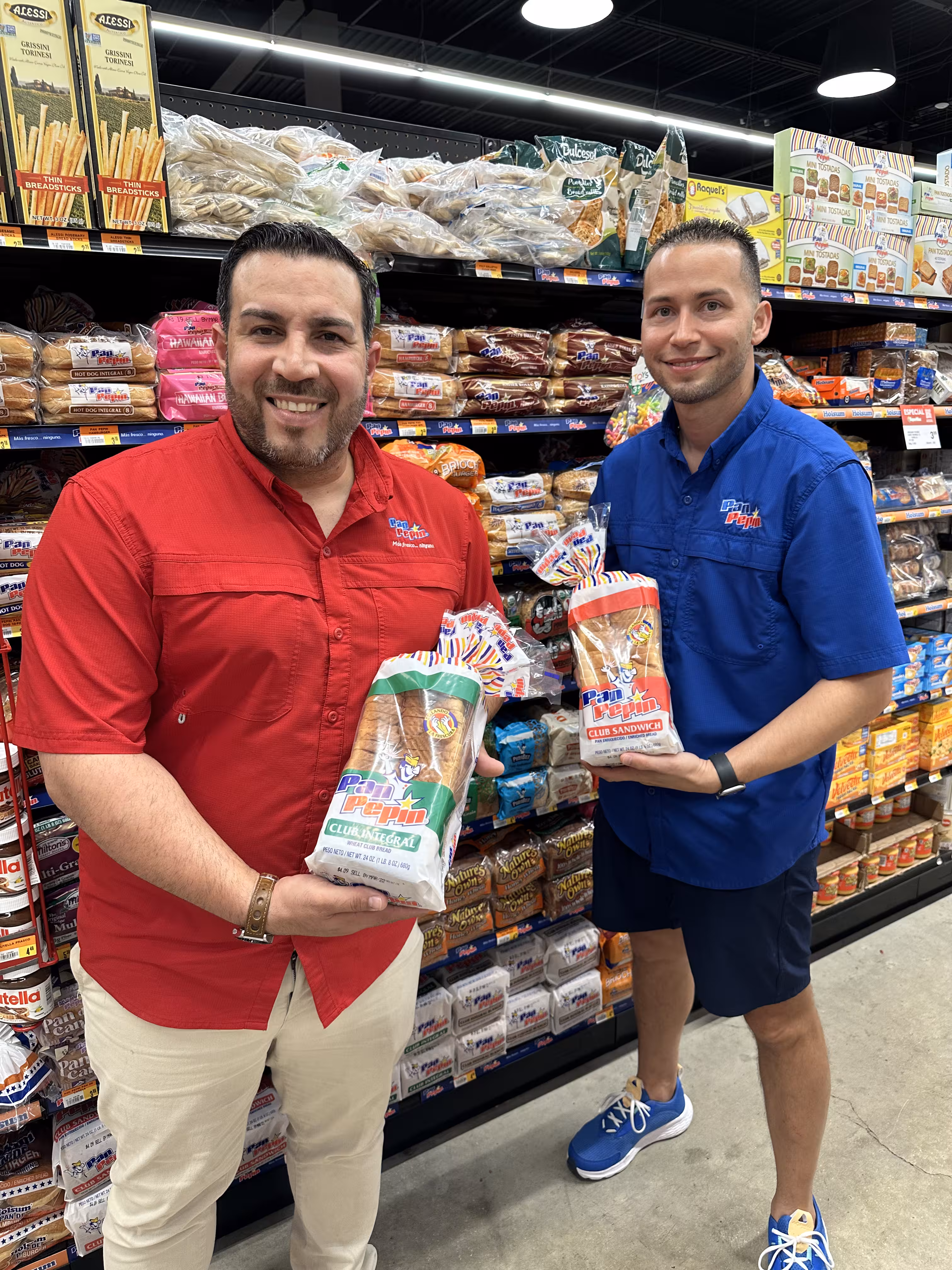 Two men in a grocery store aisle holding packages of Pan Pepin bread, one wearing a red shirt and beige pants, the other in a blue shirt and shorts.
