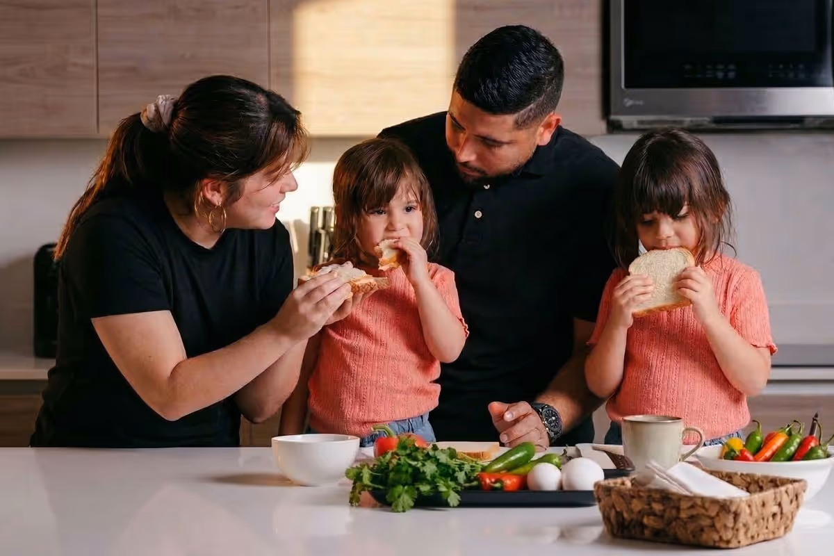 Parents and two young girls in matching pink sweaters eating bread together in a kitchen.