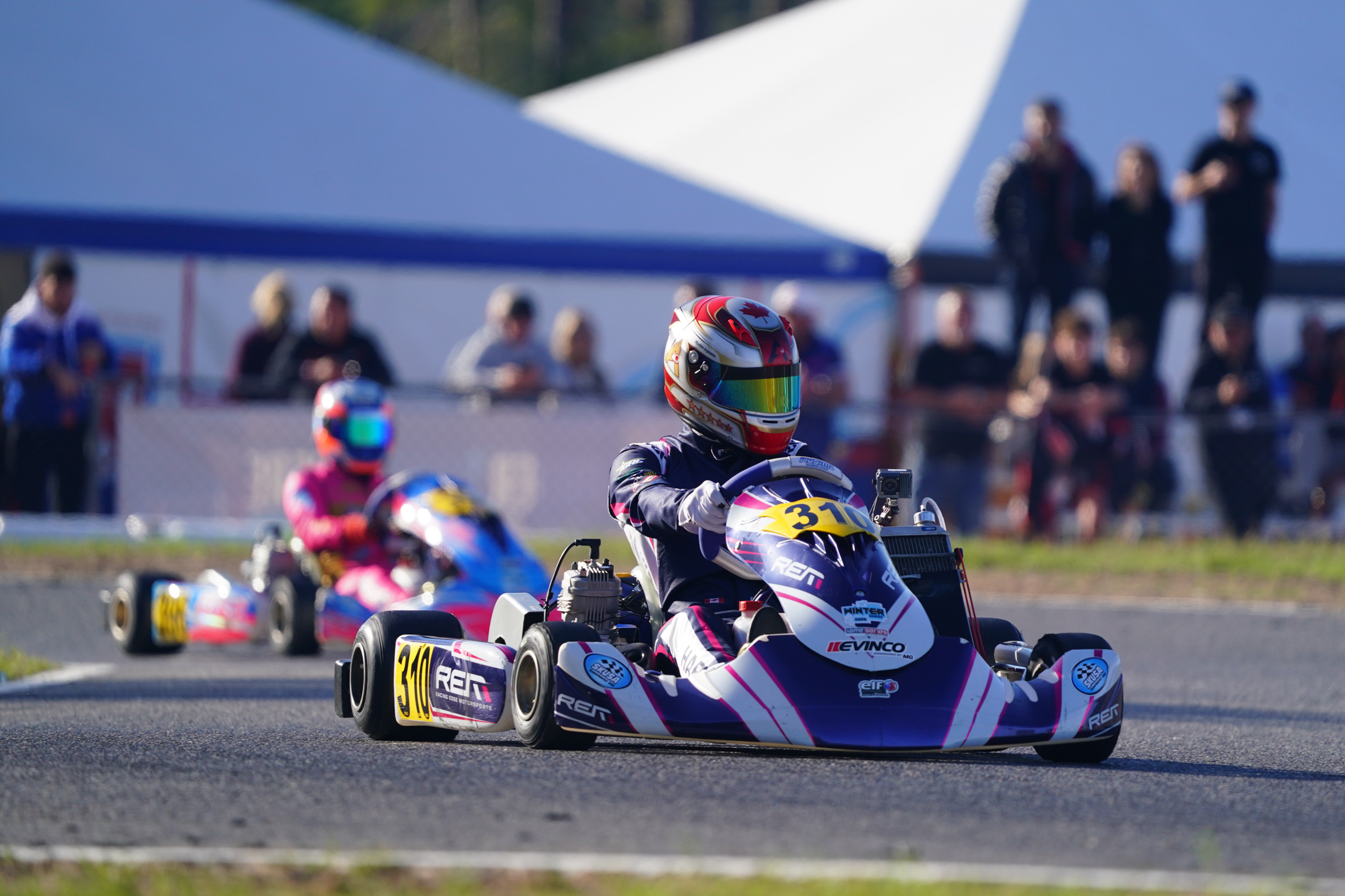 Kart racer in helmet and racing suit driving kart number 310 on a track with spectators in the background.