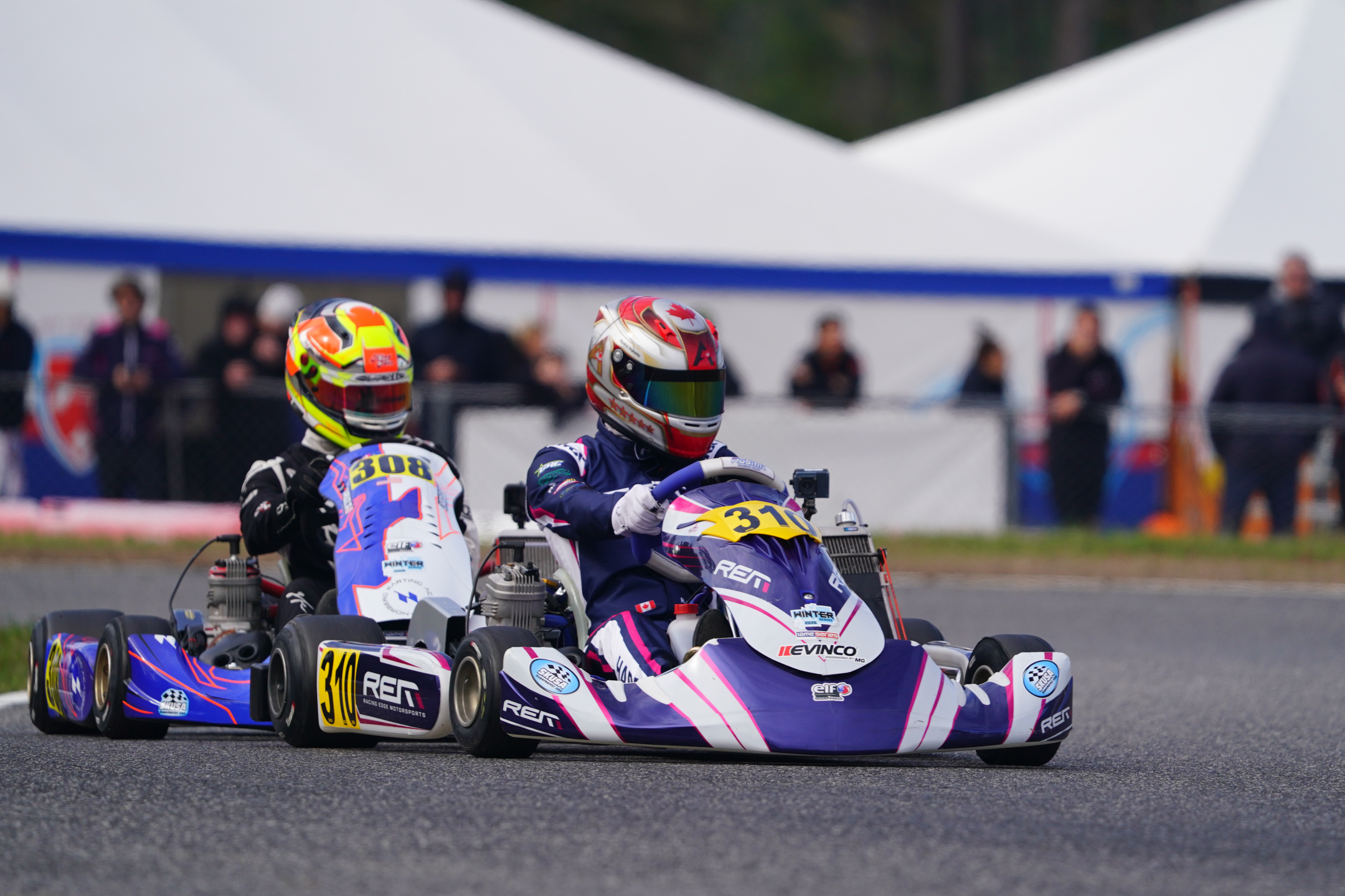 Two kart racers in helmets competing on an asphalt track with blurred spectators in the background.