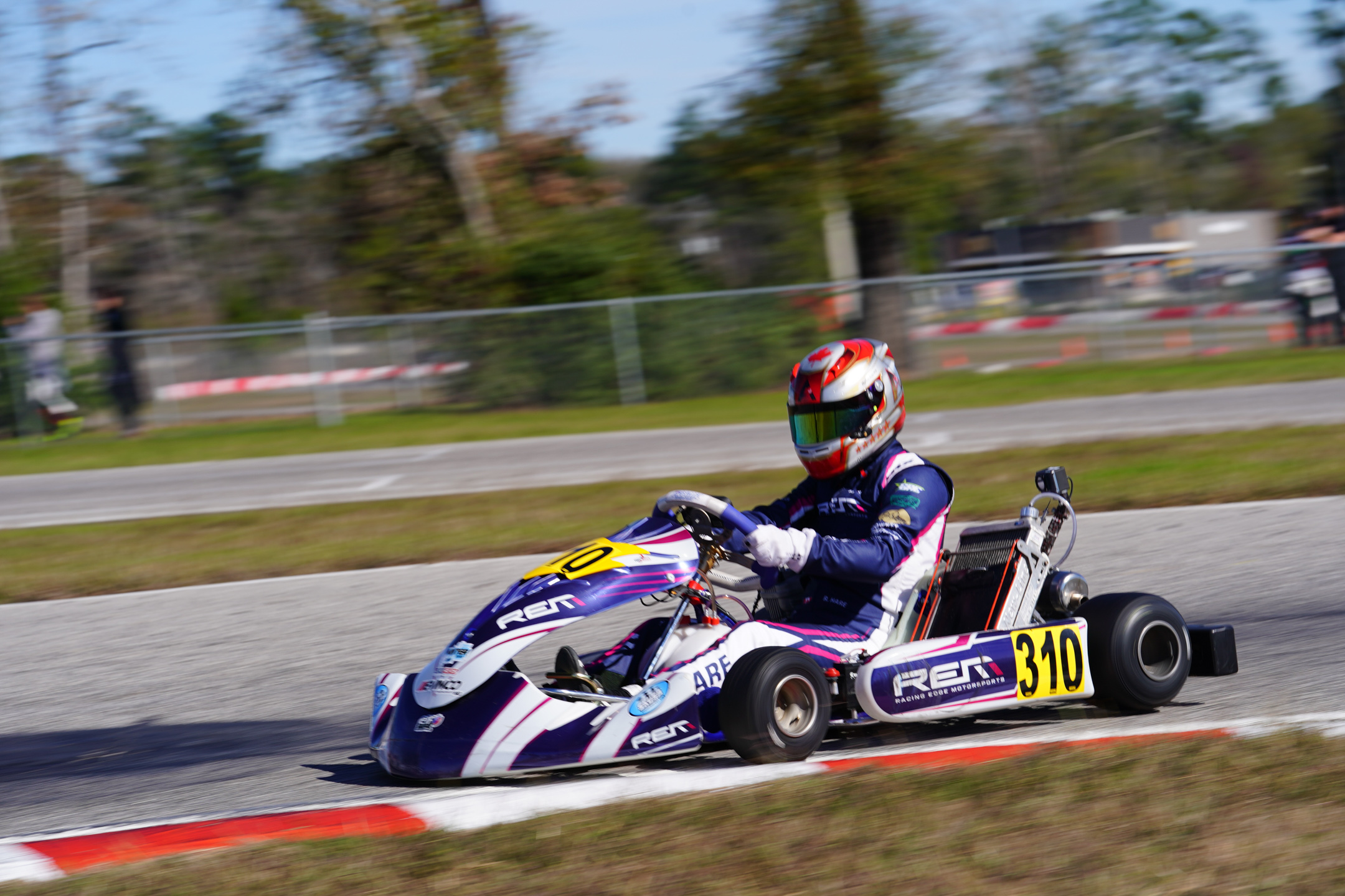 Karting driver wearing a red and white helmet racing on a track near a red and white curbing.