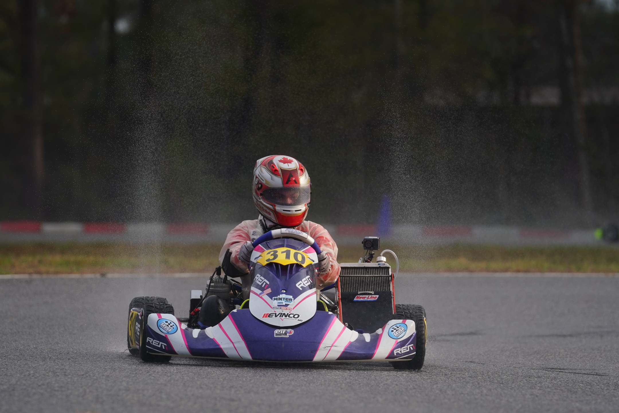 Person driving a kart with the number 310 on a wet race track, spraying water behind.