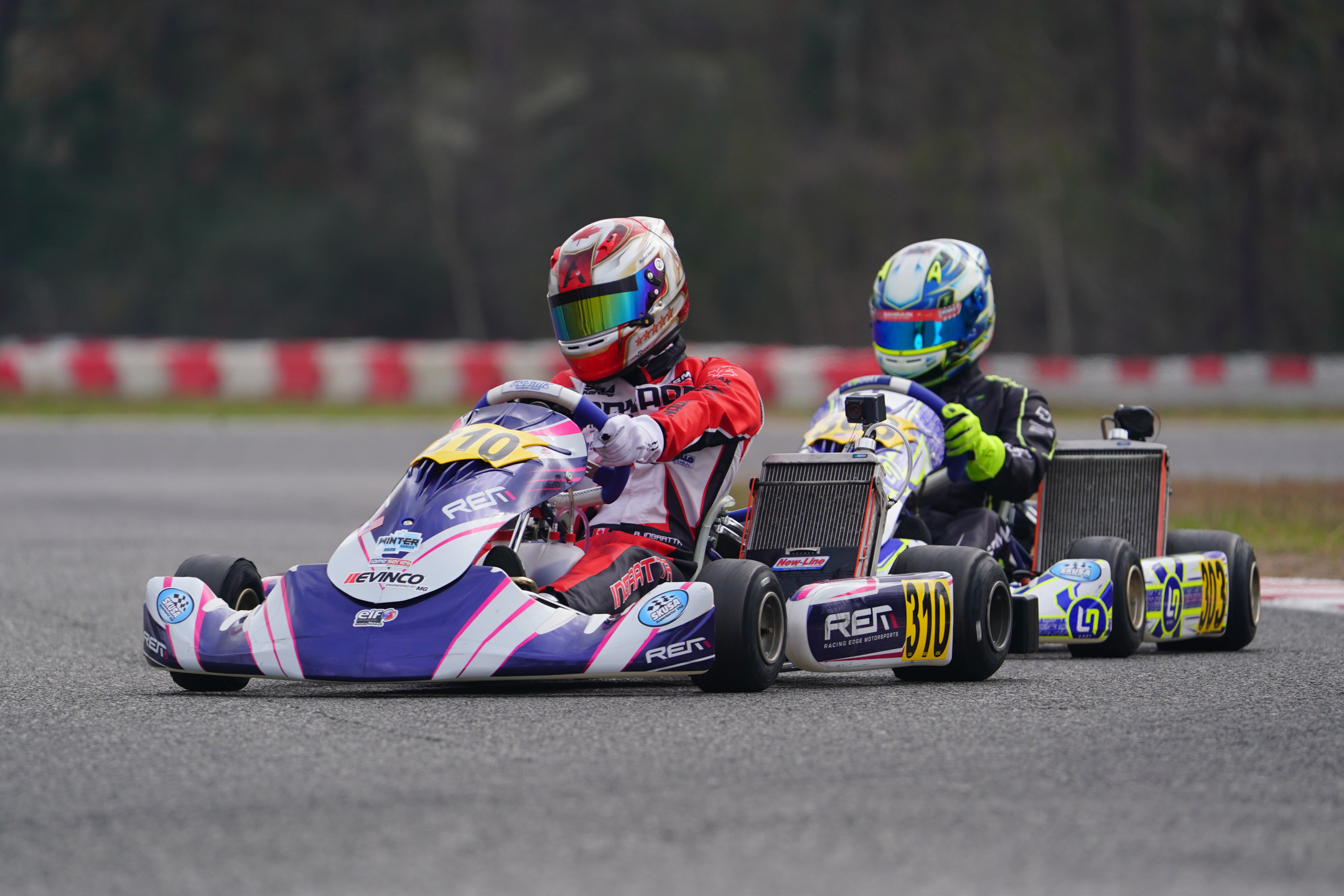 Two kart racers on a track, with the leading driver in red and white racing gear and the second driver wearing black and blue gear.