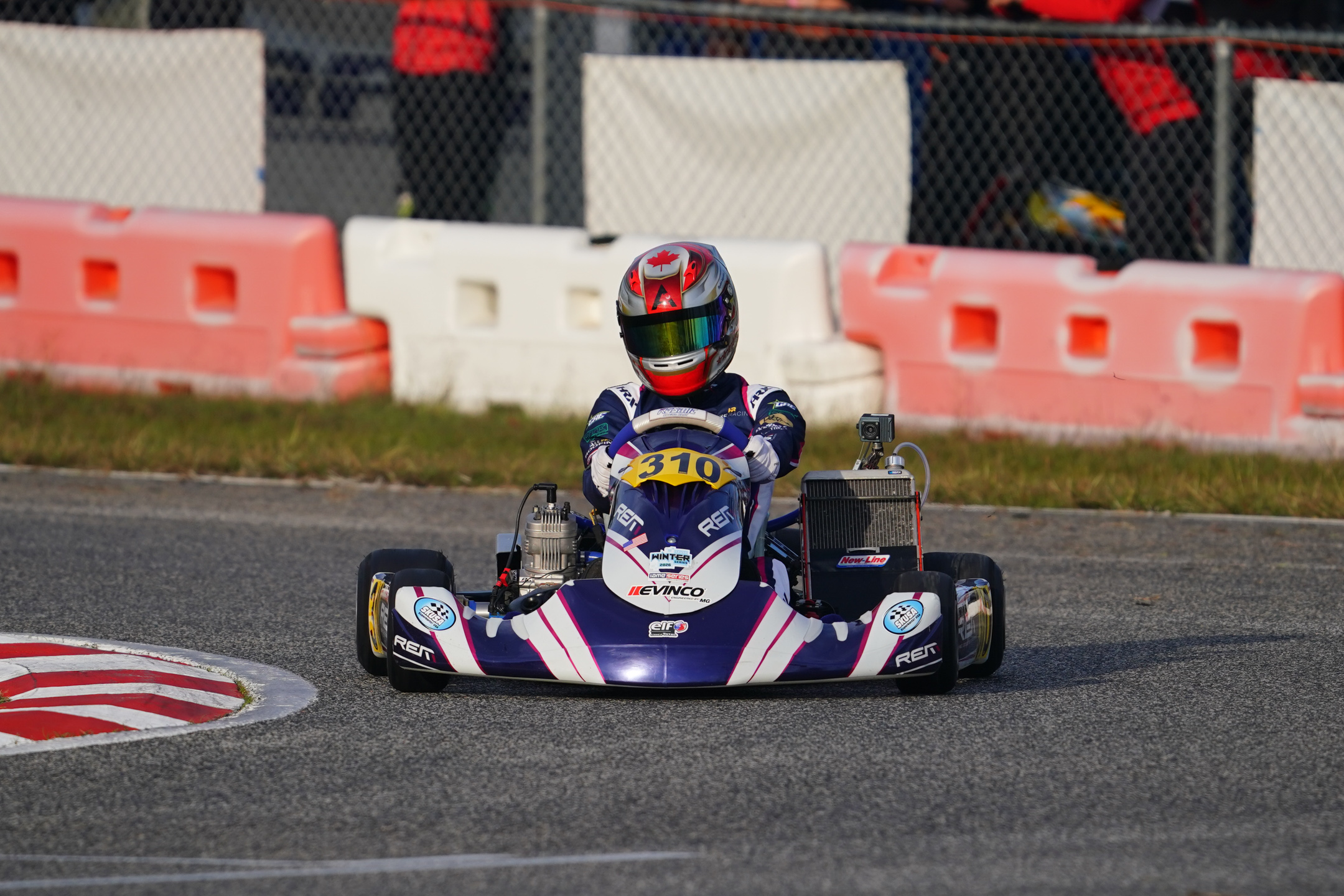 Kart racer wearing a helmet with a Canadian flag design driving a purple and white go-kart number 310 on a race track near safety barriers.