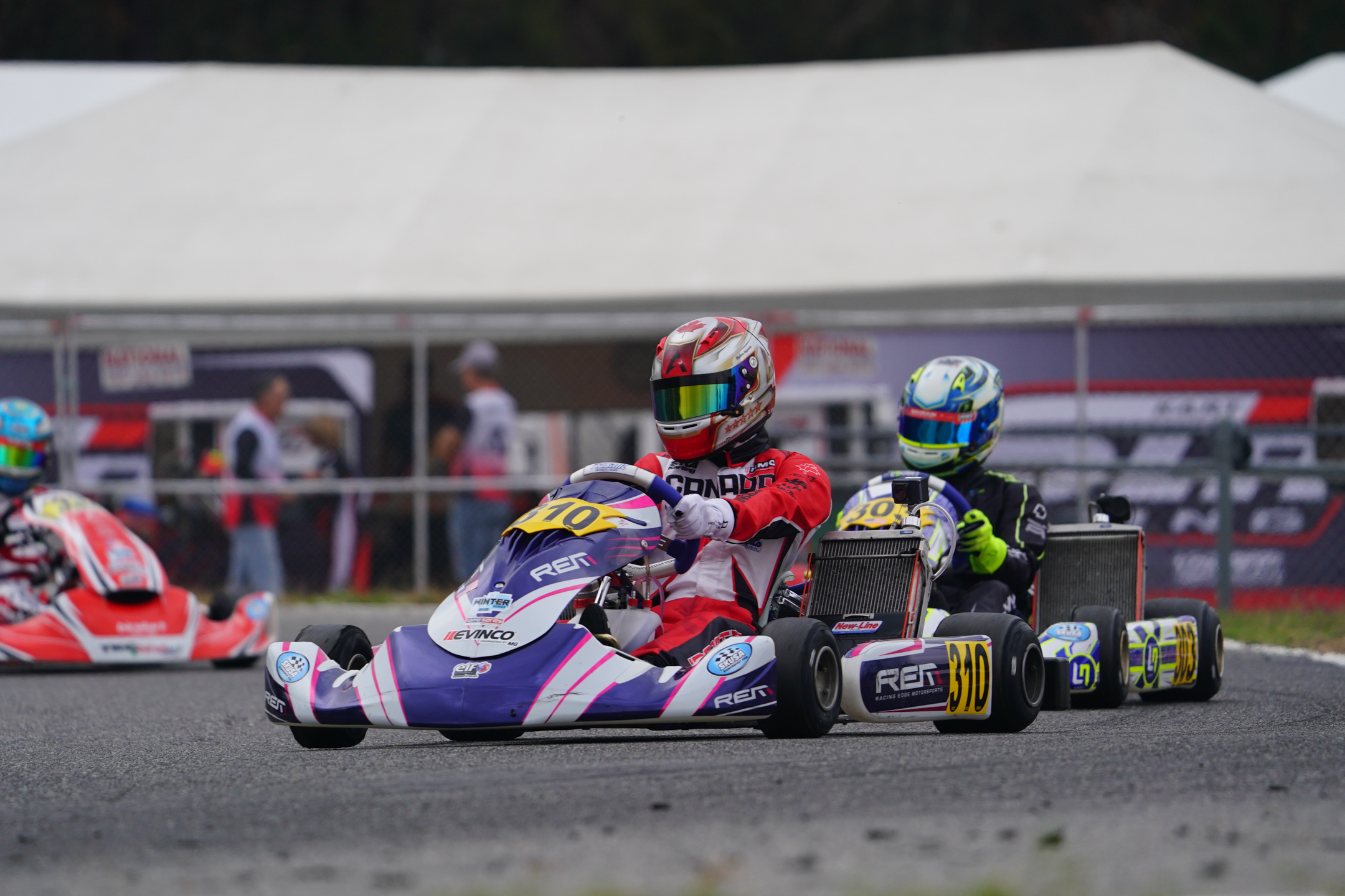 Two kart racers in helmets driving on a track, with one kart numbered 310 in front.