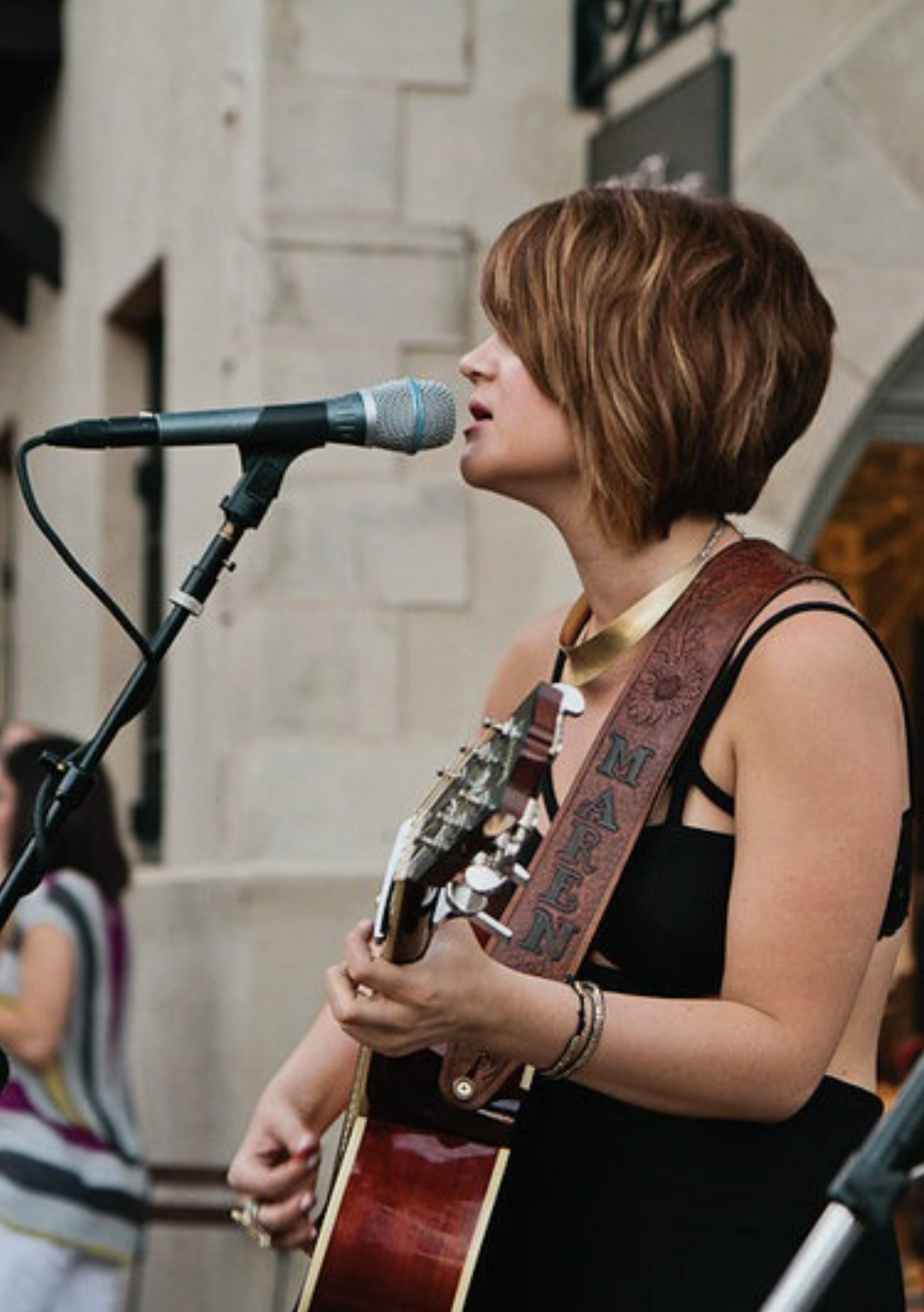 Woman with short hair singing into a microphone while playing an acoustic guitar outdoors.