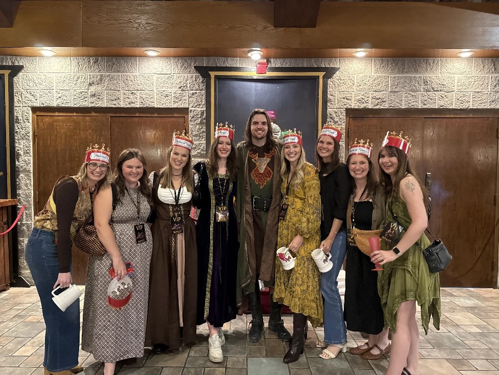 Group of eight people in medieval attire wearing red crowns posing indoors with drinks.