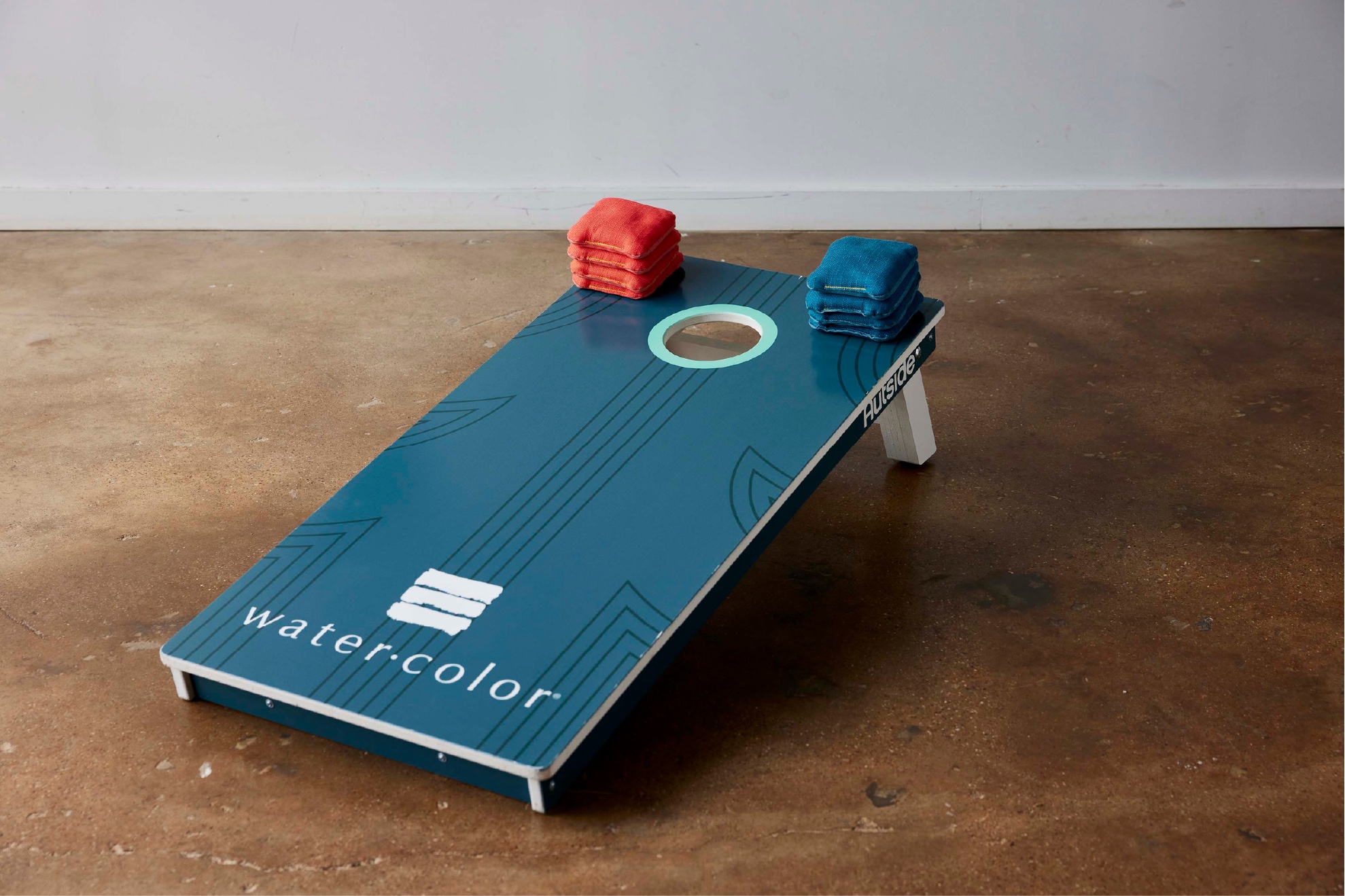 Blue cornhole board with 'watercolor' text and stacked red and blue bean bags on top.