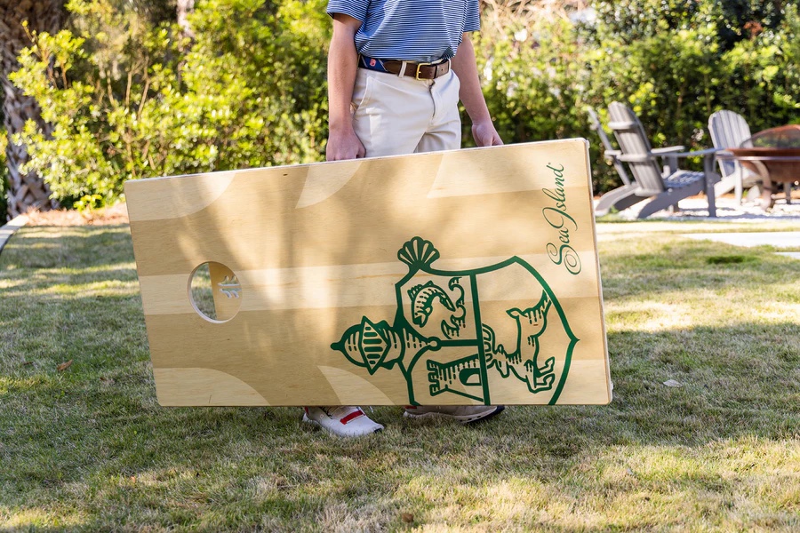 Person in blue shirt and khaki pants carrying a wooden cornhole board with Sea Island logo.