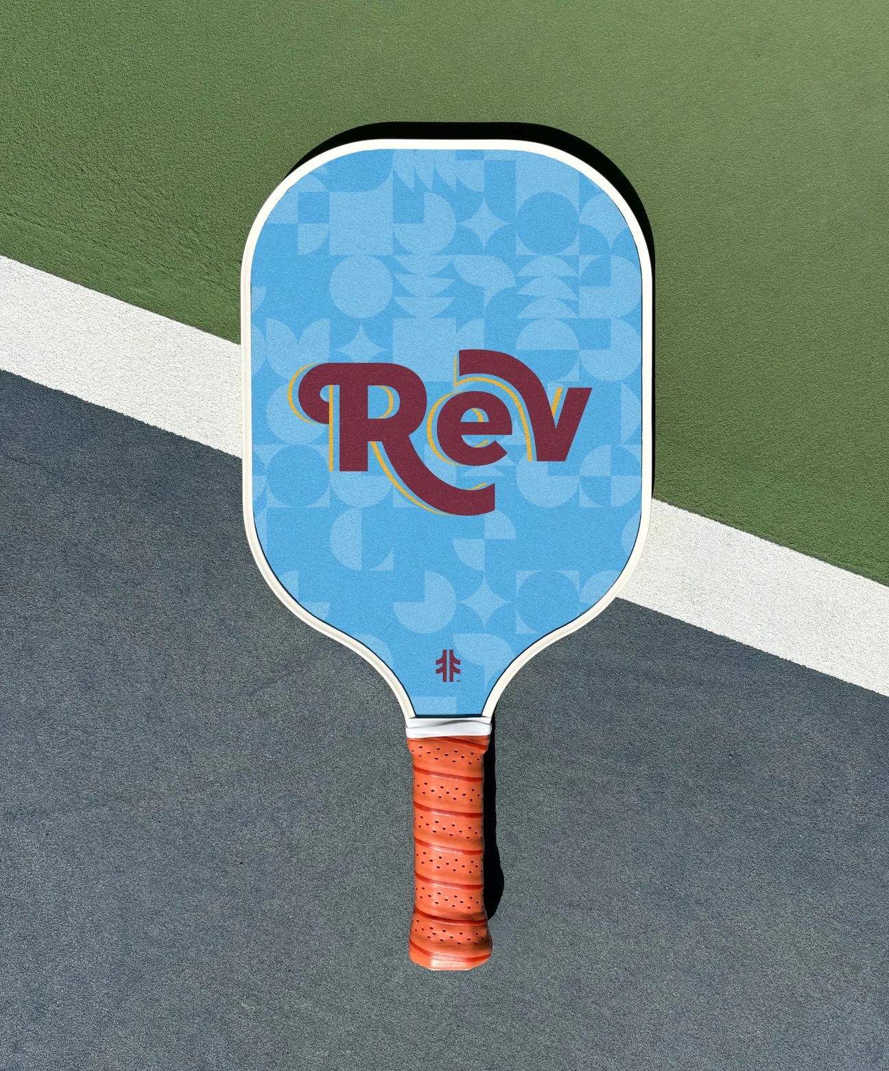 Pickleball paddle with blue face, geometric pattern, red text 'Rev,' and an orange grip on a court.