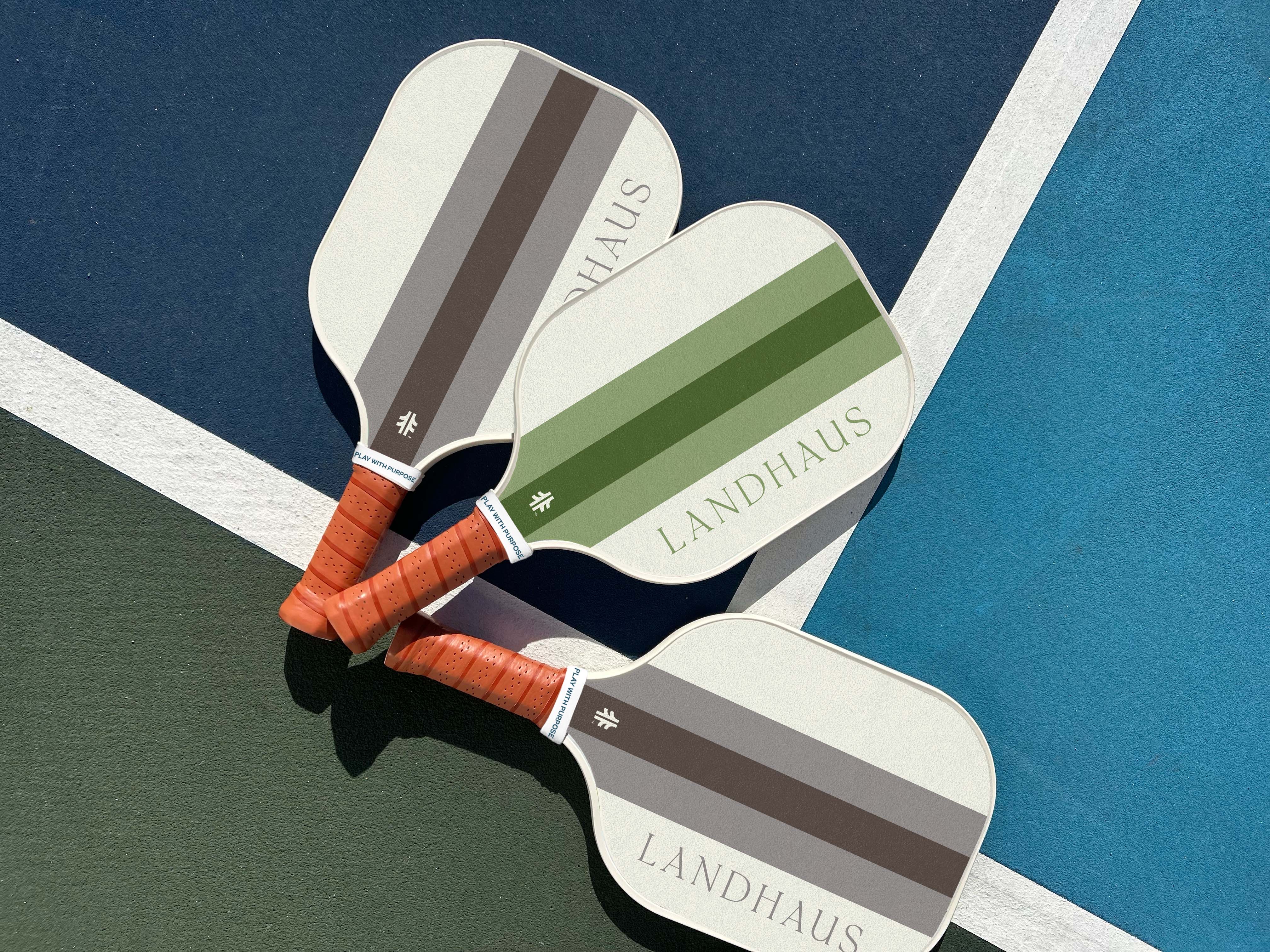 Three Landhaus pickleball paddles with brown and green stripes on a blue and green court.
