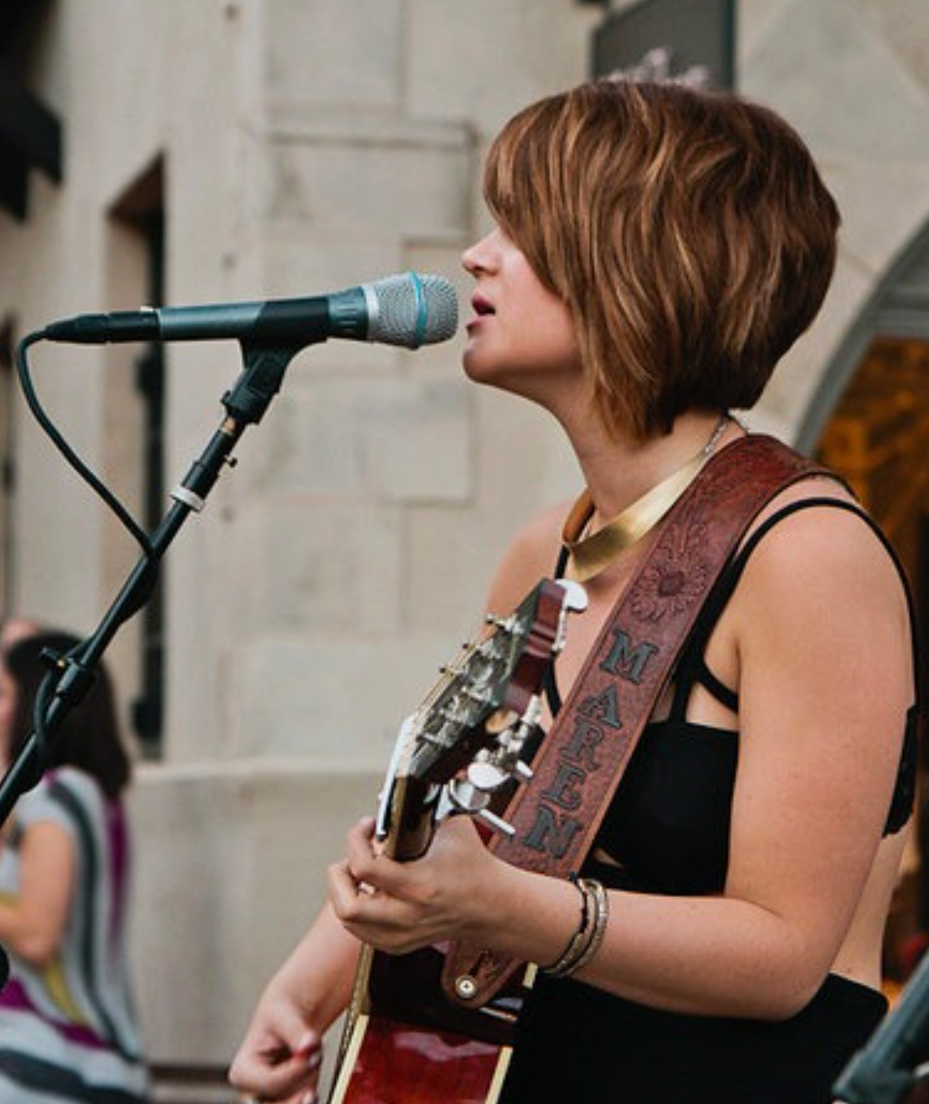 Woman singing into a microphone and playing guitar with a leather strap that says Maren.
