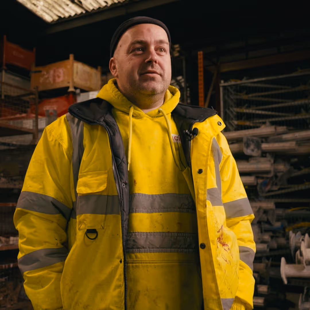 Man wearing a bright yellow reflective jacket and hoodie inside a warehouse or workshop.