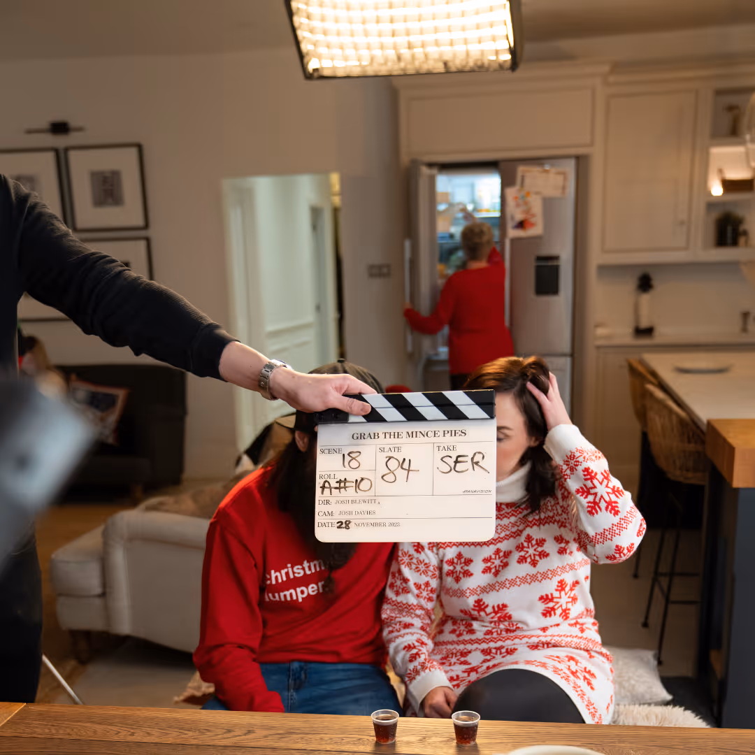 A clapperboard labeled 'Grab the Mince Pies' is held in front of two people sitting at a table, one wearing a red Christmas sweater and the other a white sweater with red snowflake patterns.