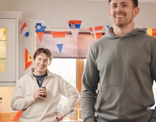 Two young men smiling indoors with international flags hung as decoration in the background.