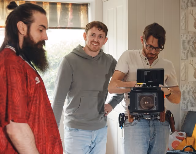 Three men indoors, one examining a professional video camera, another smiling at the camera, and the third looking down.