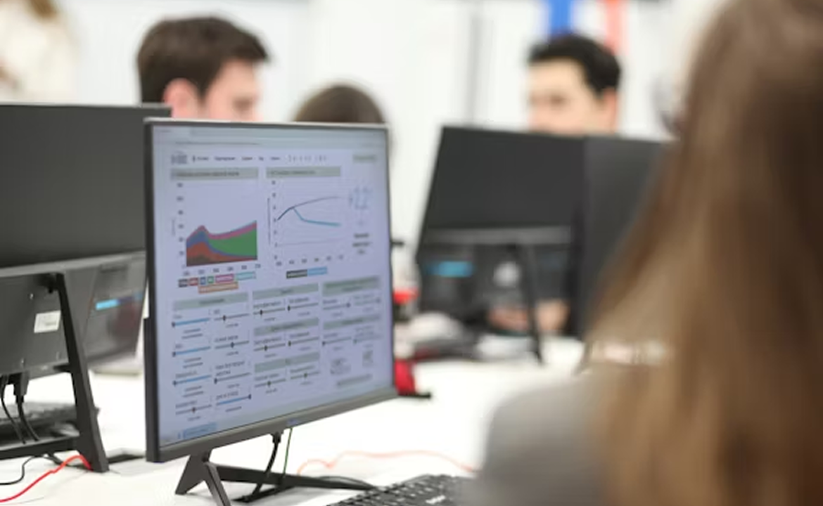 Computer monitor displaying data charts and graphs in an office setting with people working in the background.