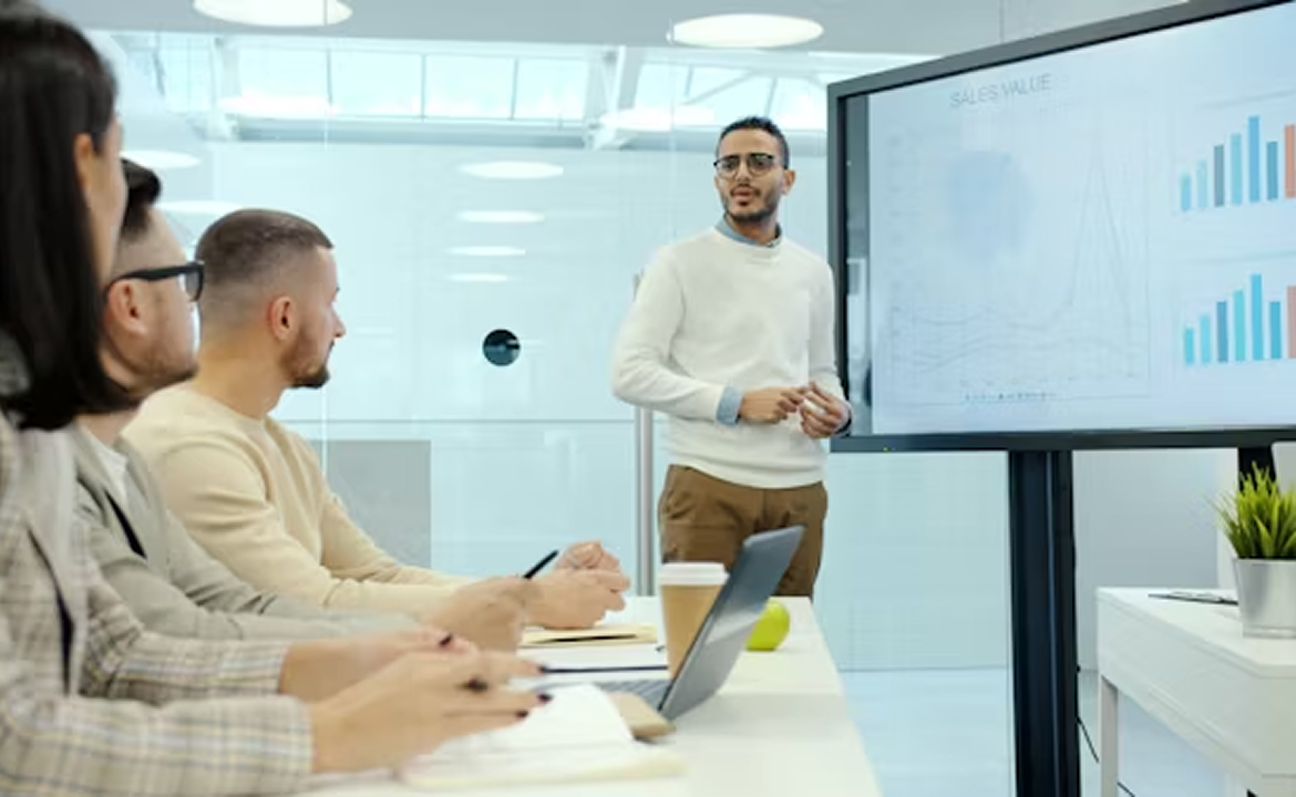 Man in white sweater giving a presentation on sales value with charts to a group of attentive colleagues in a modern office.