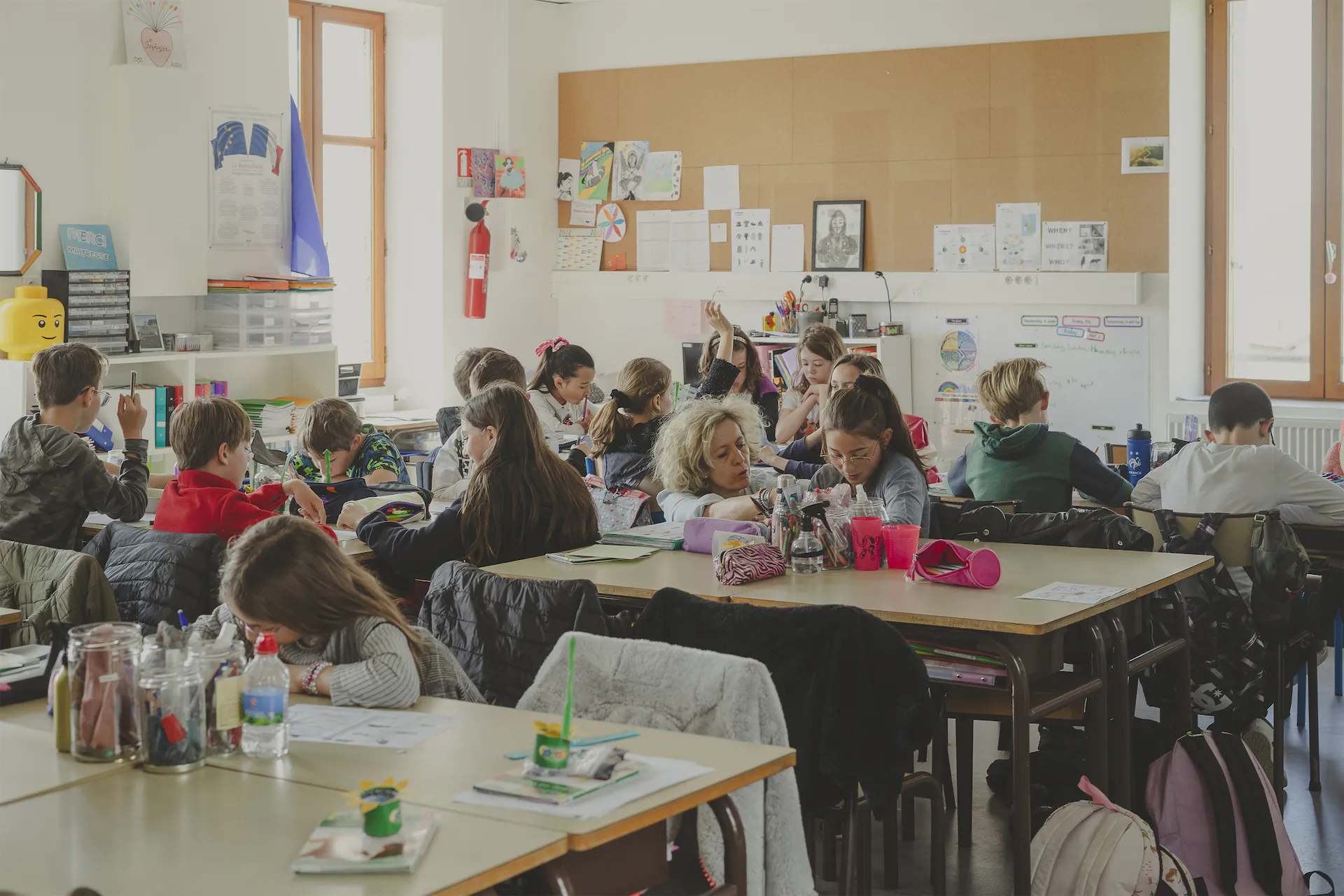 Classe de CM1-CM2 remplie d'élèves avec la maîtresse qui tourne dans la classe pour aider les élèves à réaliser l'exercice