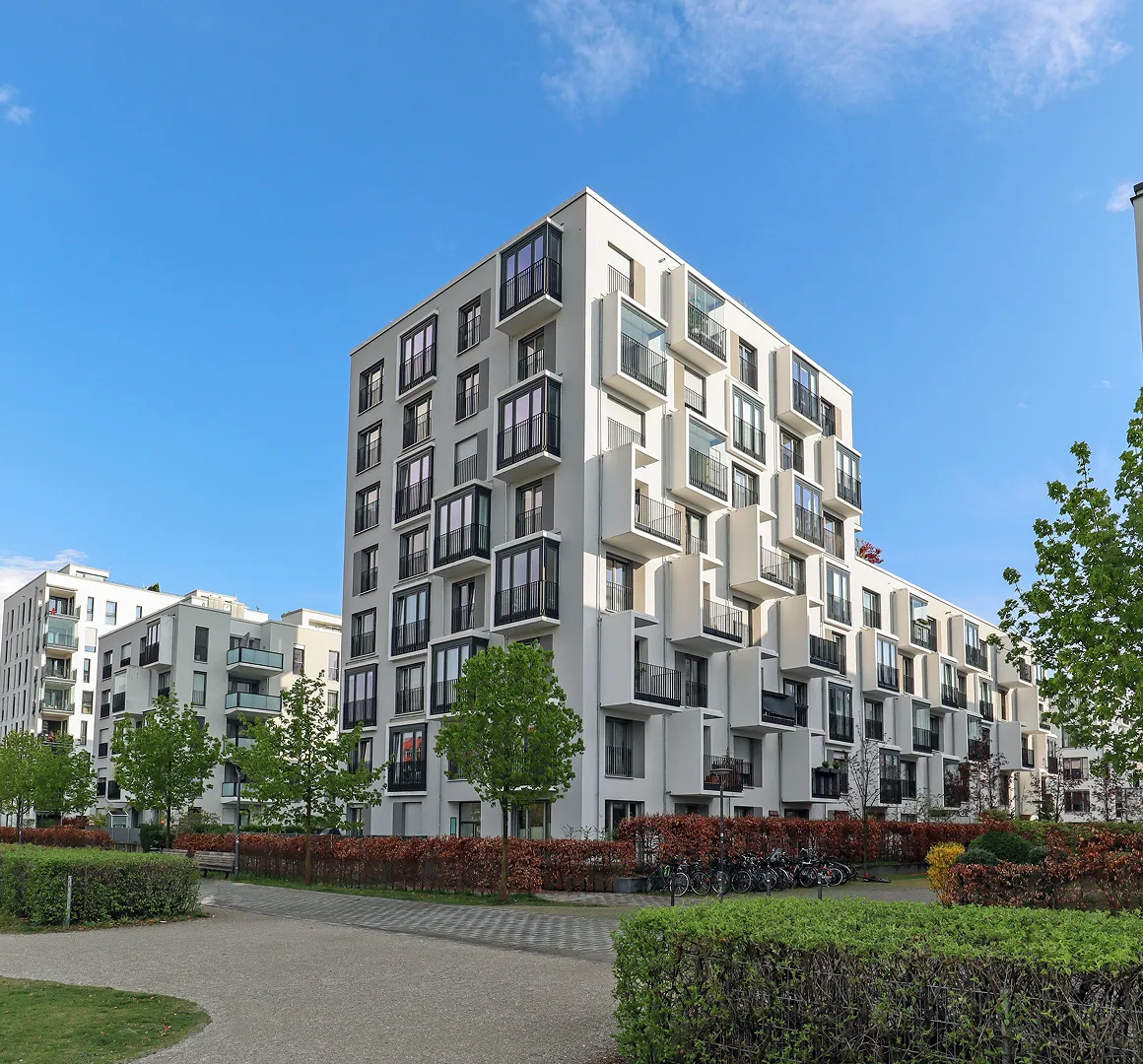 Moderne weiße Wohngebäude mit Balkonen, Bäumen und einem sonnigen blauen Himmel.