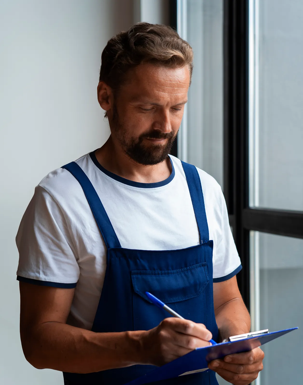 Mann in blauem Latzkleid und weißem T-Shirt schreibt auf einem Klemmbrett neben einem Fenster.