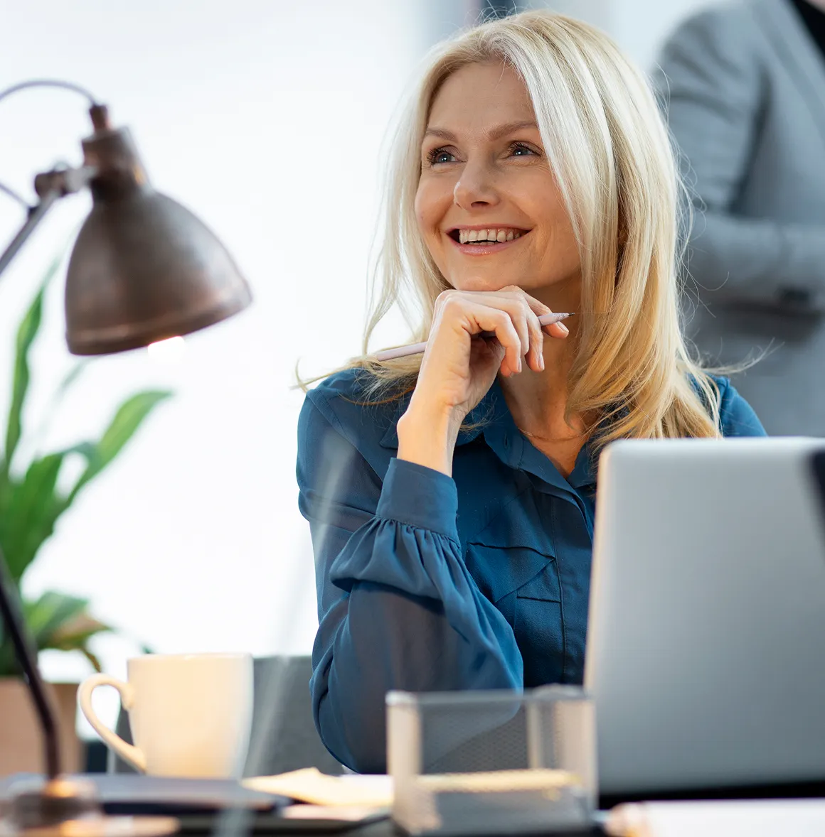 Lächelnde blonde Frau sitzt an einem Schreibtisch mit Laptop und hält einen Stift.