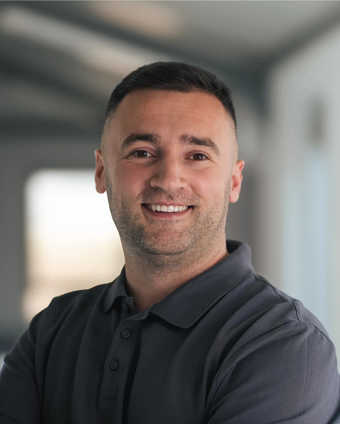 Porträt eines lächelnden Mannes mit kurzem Haar und dunklem Polohemd in einem modernen Büro.