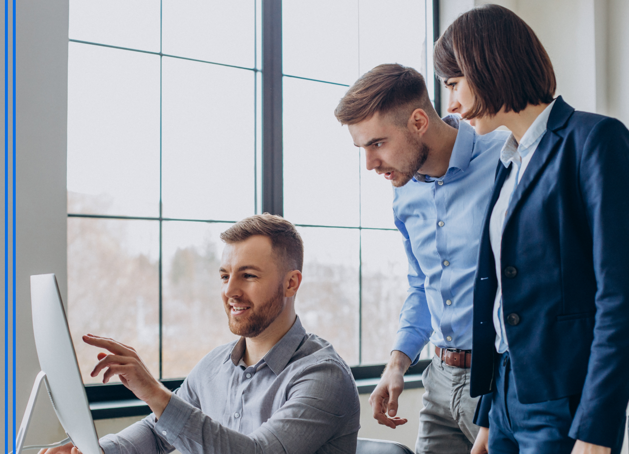 Drei junge Berufstätige diskutieren und zeigen auf einen Computerbildschirm in einem modern beleuchteten Büro.