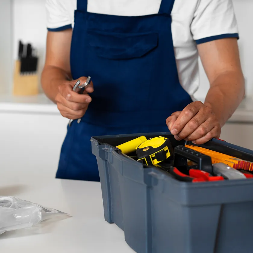 Person in blauer Schürze hält Werkzeuge über einem grauen Werkzeugkasten mit verschiedenen Mess- und Schnittwerkzeugen.