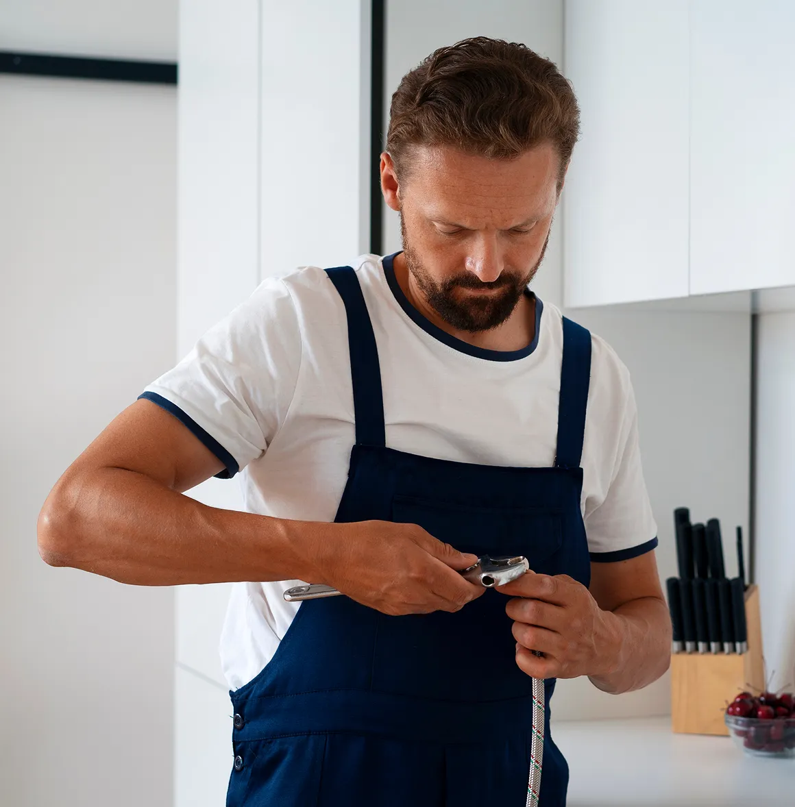 Ein Handwerker in blauer Latzhose repariert einen Schlauch mit einem Werkzeug in einer modernen Küche.